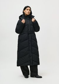 Mujer vestida con un abrigo de plumas negro largo con cinturón y capucha, sobre pantalones negros y botines negros, de pie frente a un fondo blanco liso.