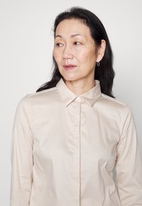 Ljust beige skjorta med krage, med en slät textur och subtil glans. Kompletteras av runda hoop-örhängen.