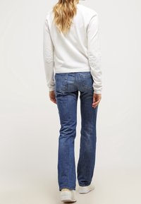 Valkoinen pitkähiha, jossa on teksturoidut kyynärpäät, yhdistettynä sinisiin liehukehousuihin ja valkoisiin lenkkareihin, nähtynä takaa.