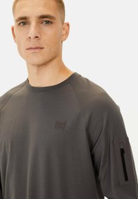Graues Langarmshirt mit rundem Ausschnitt, das eine Reißverschlusstasche am linken Ärmel und ein kleines Logo auf der Brust hat.