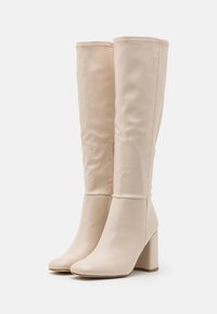 Beiga knähöga stövlar i slät syntetiskt material, med fyrkantig tå, kraftig blockklack och subtila sömnadsdetaljer.