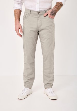 Mann trägt hellgraue Hose, weißes Hemd mit Knöpfen, weiße Sneakers, mit tätowierter linker Hand in der Tasche vor einfarbigem Hintergrund.