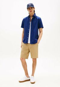 Chemise bleue à manches courtes avec boutons, shorts beige, baskets blanches avec semelles beige, et casquette marine avec un emblème. Fond uni.