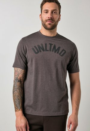 Man met baard en getatoeëerde arm die een donkergrijs T-shirt met de opdruk "UNLTMD" draagt, staand tegen een effen lichte achtergrond.
