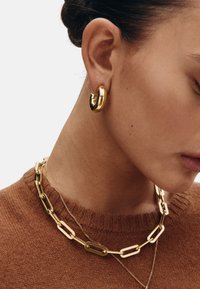 Pendiente de aro dorado y collares de cadenas doradas en capas sobre un suéter marrón. Los pendientes tienen un diseño liso y curvado; las cadenas presentan eslabones alargados.