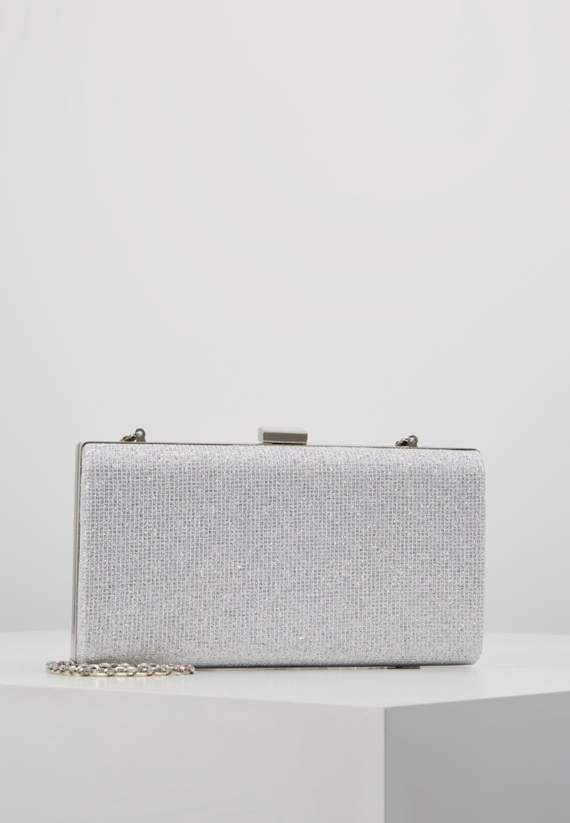 Pochette rectangulaire argentée pailletée avec cadre métallique et fermoir, posée sur une surface blanche avec une bandoulière chaîne partiellement visible.