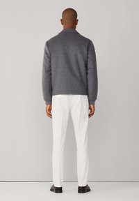 Chaqueta de lana gris con cuello, vista desde atrás. Combinada con pantalones blancos y zapatos oscuros, sobre un fondo neutro.