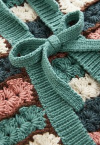 Pătură croșetată cu modele florale colorate în verde închis, roz, crem și maro, cu un detaliu texturat și cu fundiță din lână verde deschis.