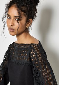 Czarna odzież z misternymi koronkowymi detalami na rękawach, o gładkiej teksturze i fasonie odsłaniającym ramiona. Złote kolczyki podkreślają wygląd.