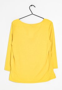 Żółta bluzka z długim rękawem wisząca na czarnym wieszaku na białym tle, pokazująca widok z tyłu z okrągłym dekoltem i luźnym krojem.