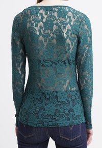 Tealfärgad spetsblus med långa ärmar som har blommönster, semi-transparent textur och rund halsringning. Ställd i kombination med mörka denimjeans.