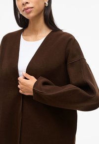 Rudas, ilgos rankovės kardiganas iš minkštos medžiagos su laisvu siluetu ir giliu V formos iškirpimu, dėvimas ant balto marškinėlio, su matomomis siūlėmis.