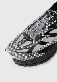 Chaussure de course avec une tige en maille perforée noire et blanche avec des motifs dynamiques, des lacets ronds et une semelle en caoutchouc noire rembourrée.