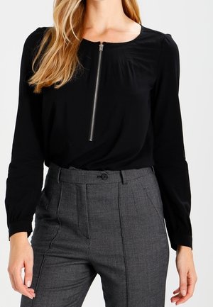 Zwart shirt met lange mouwen, een ronde halslijn en ritssluiting aan de voorkant, gecombineerd met hooggetailleerde grijze geruite broek met plooien.