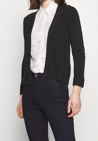 Czarny dzianinowy kardigan z otwartym przodem, ściągaczami na mankietach i u dołu. Noszony na jasnej, wzorzystej bluzce. Do kompletu ciemne dżinsy.