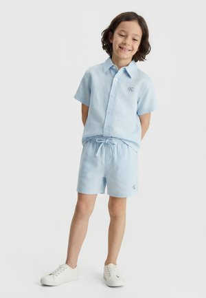 Kind lächelt, steht mit den Händen hinter dem Rücken, trägt ein hellblaues kurzärmliges Hemd mit Knöpfen, passende Shorts und weiße Sneakers vor weißem Hintergrund.