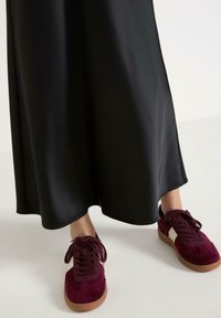 Schwarzer Satin-Maxirock mit glatter Textur, kombiniert mit burgunderfarbenen Wildledersneakern mit Schnürung und hellbraunen Gummisohlen.