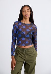 Dolga majica iz prozorne črne mreže s modrim cvetličnim vzorcem, z okroglo izrezom in krajšo obliko. V kombinaciji z olivno zelenimi hlačami.