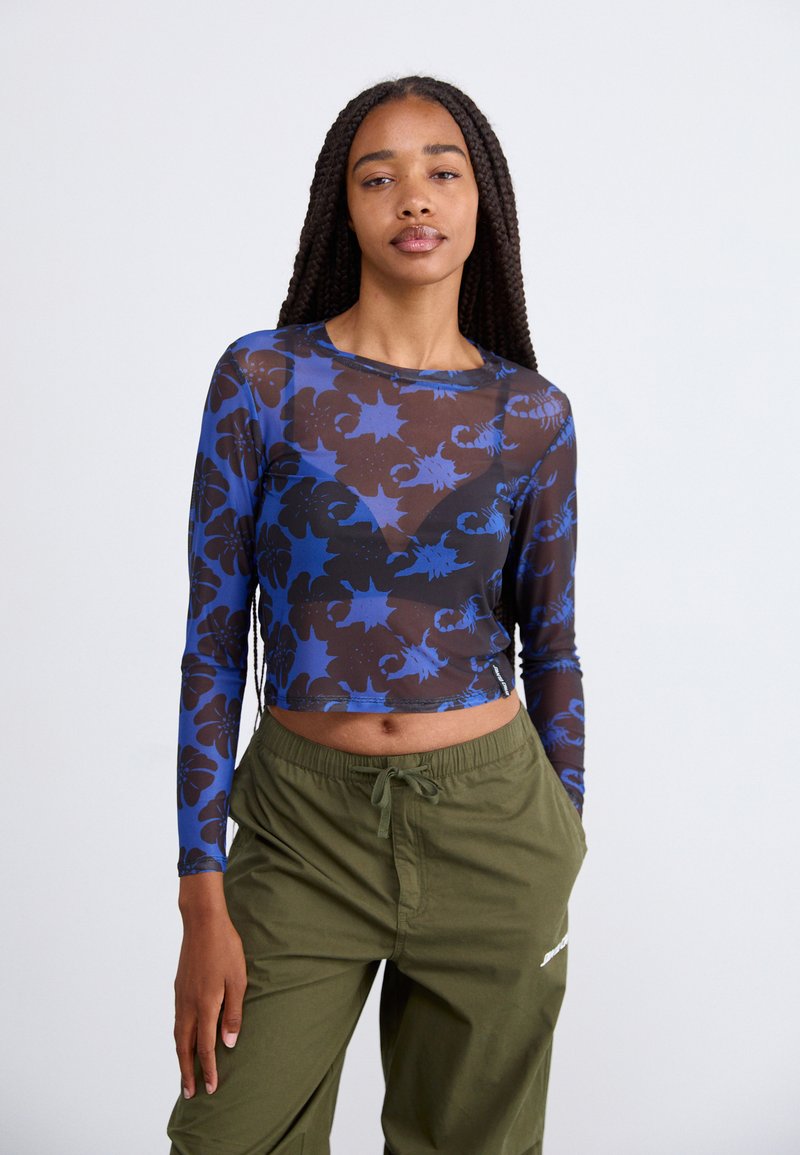 Dolga majica iz prozorne črne mreže s modrim cvetličnim vzorcem, z okroglo izrezom in krajšo obliko. V kombinaciji z olivno zelenimi hlačami.