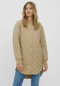 Manteau beige matelassé avec un devant à boutons, des manches longues, des zips sur les côtés et un ourlet arrière légèrement plus long, associé à un jean bleu.