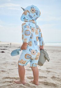 Light blue sun suit with animal cartoon patterns, long sleeves, and a hood. Child holding a green bucket and blue shovel on sandy beach.
