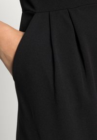 Hand partially inside the pocket of a black pleated garment, highlighting fabric texture and pocket design.