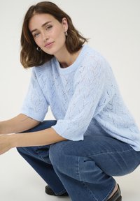 Gaiši zils adīts džemperis ar īsām piedurknēm un atvērtu audumu; saskaņots ar ziliem džinsu biksēm; modele noliecusies pret neitrālu fonu.