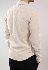 Beige kordfløyelskjorte med lange ermer, knapper på mansjettene og buet nederkant. Har et lite oransje logo nær bunnen. Teksturert ribbet stoff.