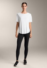 Bílé tričko s krátkým rukávem a zakulaceným lemem, v kombinaci s černými legínami a černými sportovními botami na neutrálním pozadí.