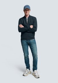 Schwarze Bomberjacke mit Reißverschluss, weißes T-Shirt, blaue Jeans und weiße Sneaker. Eine graue Mütze rundet das lässige Outfit ab.