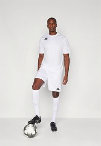 Equipación deportiva blanca que consiste en una camiseta de manga corta y pantalones cortos, con detalles de logotipo en negro, combinada con tacos negros y un balón de fútbol con diseño estampado.