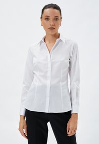 Biała koszula z długim rękawem, zapinana na guziki, wykonana z gładkiego materiału, z klasycznym kołnierzykiem, dopasowanym krojem i subtelnym detalem szycia wzdłuż szwów.