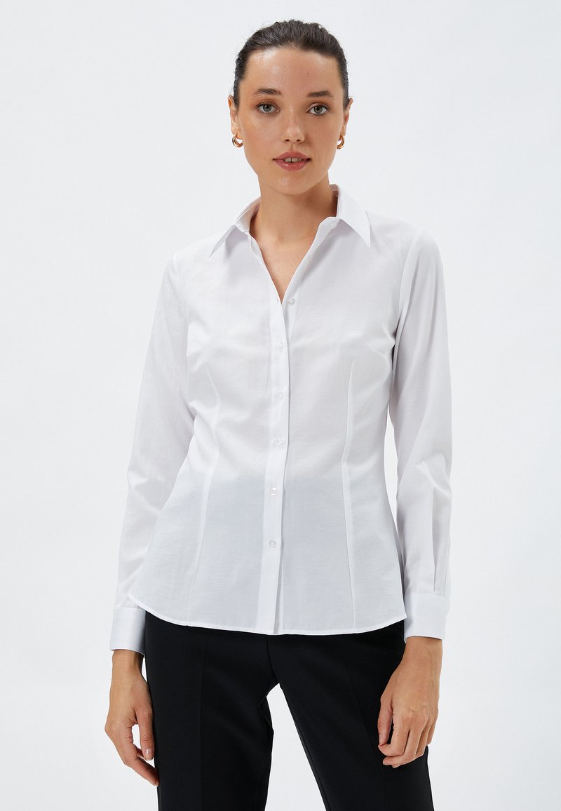 Biała koszula z długim rękawem, zapinana na guziki, wykonana z gładkiego materiału, z klasycznym kołnierzykiem, dopasowanym krojem i subtelnym detalem szycia wzdłuż szwów.