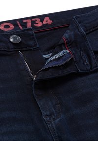 Jeans de color azul oscuro con cierre de cremallera, botón de cierre y costuras rojas. Presenta una etiqueta con el número "1734" visible en la cintura.