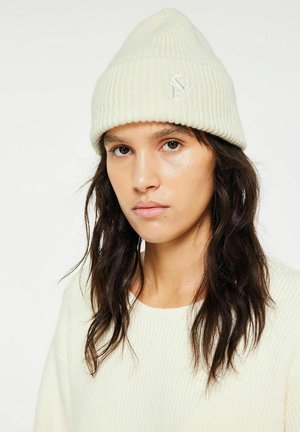 Joven mujer con cabello largo y oscuro, que lleva un gorro de punto color crema y un suéter a juego, mirando al frente con una expresión neutra.