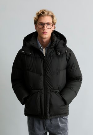 Jeune homme aux cheveux blonds bouclés et portant des lunettes, vêtu d'une doudoune noire à capuche, les mains dans les poches, sur fond uni.