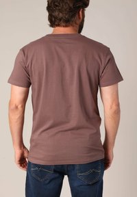 Man met kort bruin haar, gekleed in een effen bruin T-shirt met korte mouwen en blauwe jeans, met zijn rug naar de camera gericht tegen een neutrale achtergrond.