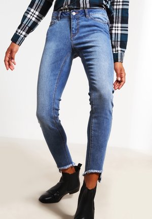 Slim-fit blauwe jeans met versleten zomen, gedragen met zwarte enkellaarzen en een blauw-wit geruit overhemd.