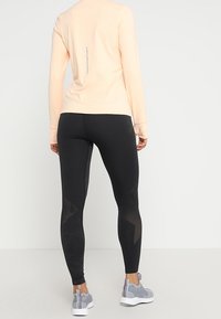 Jasnopomarańczowa bluzka z długim rękawem i szwem z tyłu, w połączeniu z czarnymi legginsami z siateczkowymi akcentami oraz szarymi butami sportowymi z fakturowanymi podeszwami.