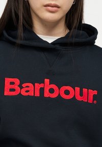 Inimese lähivõte musta kapuutsiga, rinnal suur punane tekst "Barbour", näidates suu ja kaela detaile.