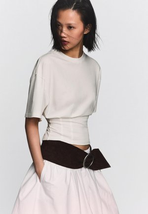 Ung kvinde med kort sort hår, iført en cremefarvet tætsiddende top, bredt mørkt bælte med metalspænde og hvid nederdel, hænderne i lommerne, foran en ensfarvet baggrund.