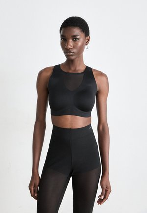 Kvinne som står mot en hvit bakgrunn iført en svart ermeløs croptopp og høyt liv, gjennomsiktige svarte leggings.