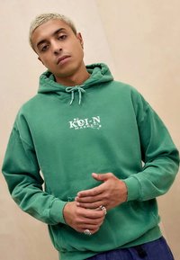 Grön bomulls-hoodie med framficka, justerbar huva med snöre och vit "KOLN MARKET"-logotyp. Har en avslappnad passform och mjuk textur.