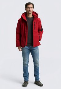 Červená prešívaná bunda s kapucňou, predným zipsom a bočnými vreckami; nosená cez sivý sveter a modré džínsy; gumové topánky s podrážkou.