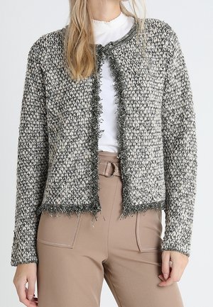 Femme portant un cardigan gris et blanc texturé à ouverture sur le devant avec des bords à franges, un chemisier blanc et un pantalon beige ceinturé.
