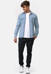 Joven de pie, vistiendo una camisa de mezclilla azul claro sobreuna camiseta blanca, pantalones negros y zapatillas blancas, sonriendo y mirando hacia abajo.