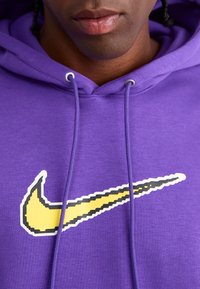 Violetinė pūkinė striukė su dideliu, pikseliuotu geltonu ir juodu Nike swoosh logotipu. Yra raišteliai ir surištos rankovės. Minkšta audinio tekstūra.