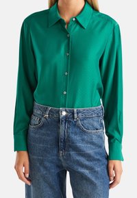 Grön skjorta med minut prickmönster, långa ärmar och krage, i kombination med blå högmidjade jeans.