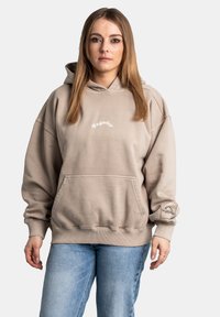 Sudadera oversized beige con un bolsillo frontal, puños acanalados y logotipo bordado "Pegador" en el pecho y la manga. Diseño casual.