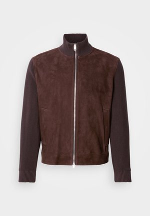 Brązowa zamszowa kurtka z prążkowanymi rękawami. Posiada wysoki kołnierz i przedni zamek błyskawiczny, boczne kieszenie oraz gładką teksturę.